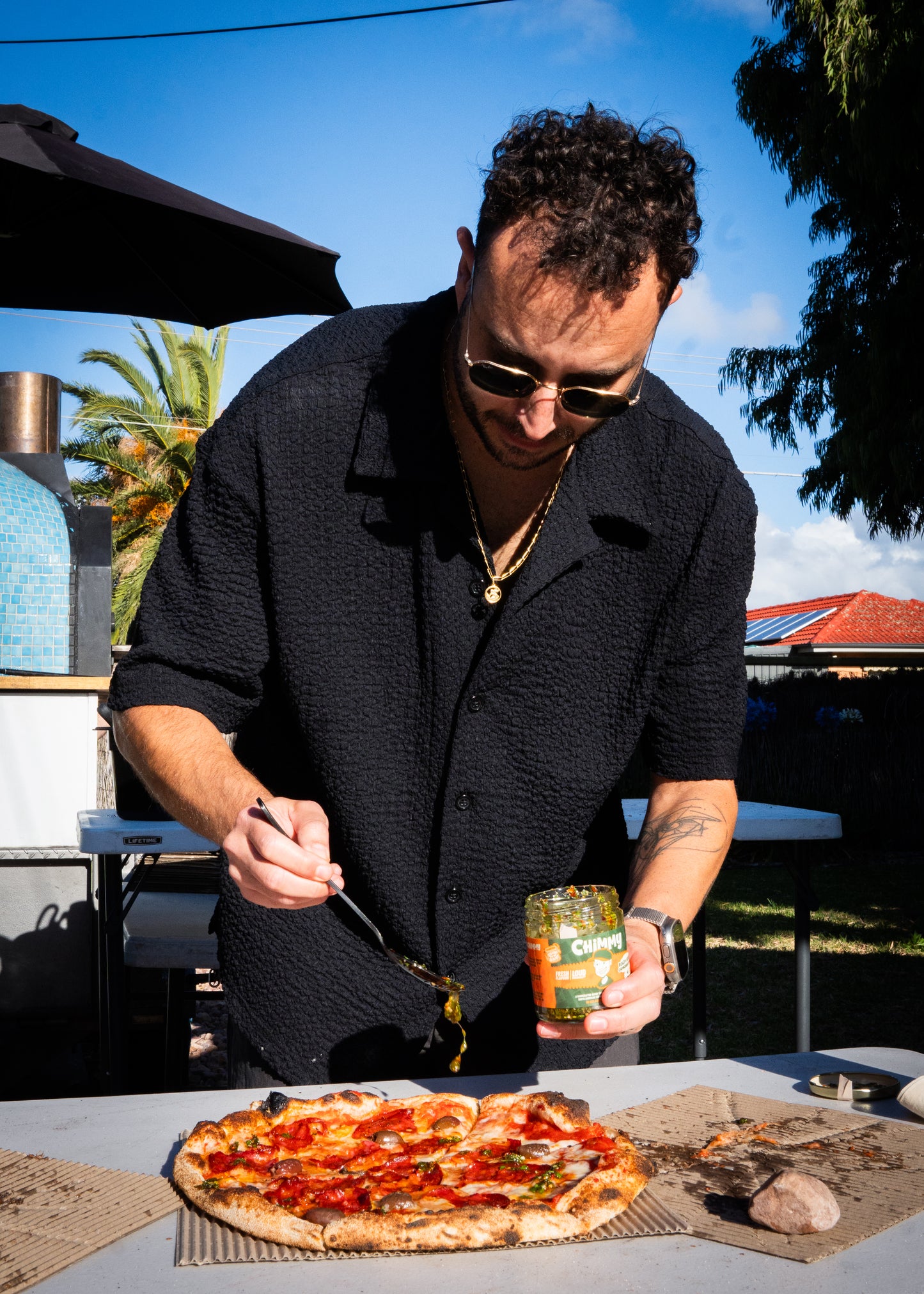 Person spooning Chimmy chimichurri onto a wood-fired pizza beside an outdoor pizza oven
