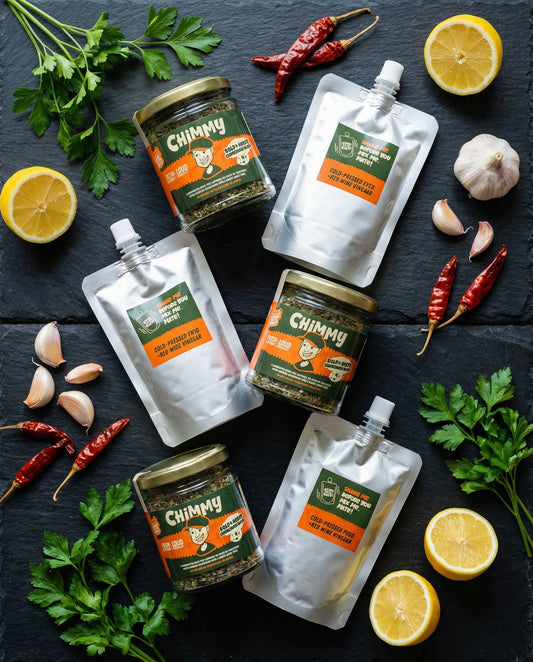Three Chimmy chimichurri kits styled with fresh parsley, lemons, garlic and dried chillies on a slate background