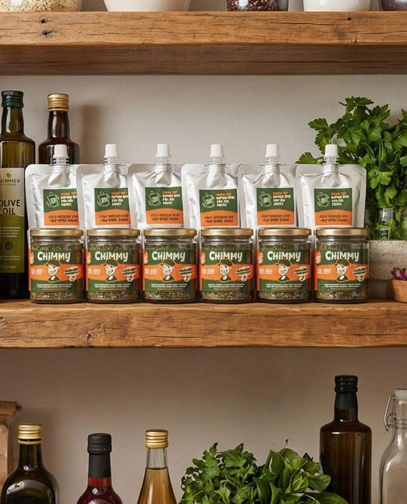 Six Chimmy chimichurri kits lined up on a wooden kitchen shelf alongside olive oil bottles and fresh herbs