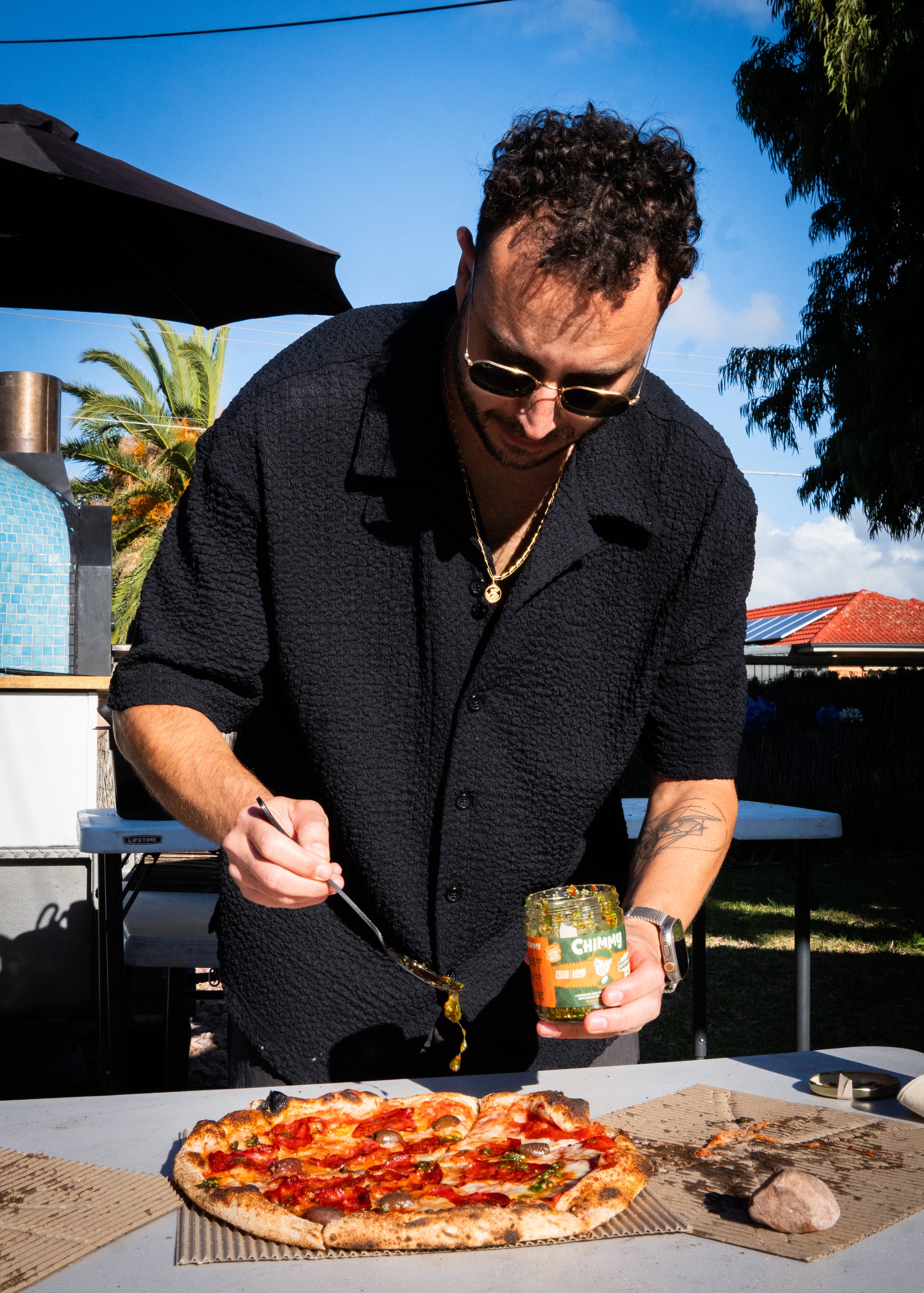 Person spooning Chimmy chimichurri onto a wood-fired pizza beside an outdoor pizza oven