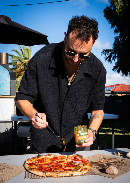 Person spooning Chimmy chimichurri onto a wood-fired pizza beside an outdoor pizza oven