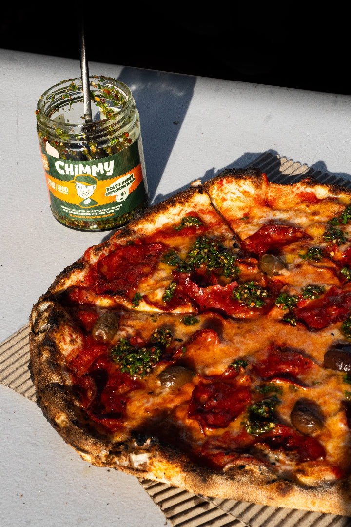 Wood-fired pizza topped with Chimmy chimichurri sauce next to an open Chimmy herb jar